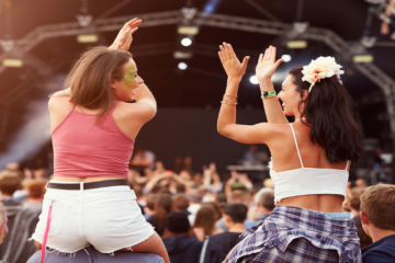 Musikfestivals 2024- Zwei junge Frauen sitzen auf den Schultern von Freunden lachend an einem Konzert.